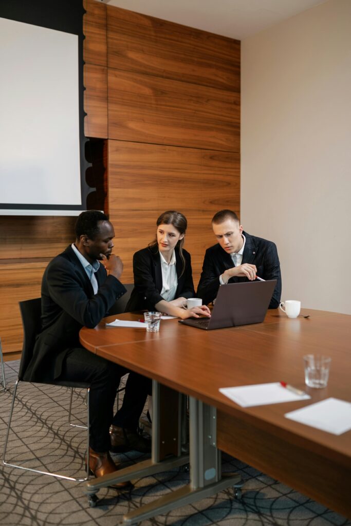 Three colleagues engaged in a business discussion around a wooden table in a contemporary conference room.